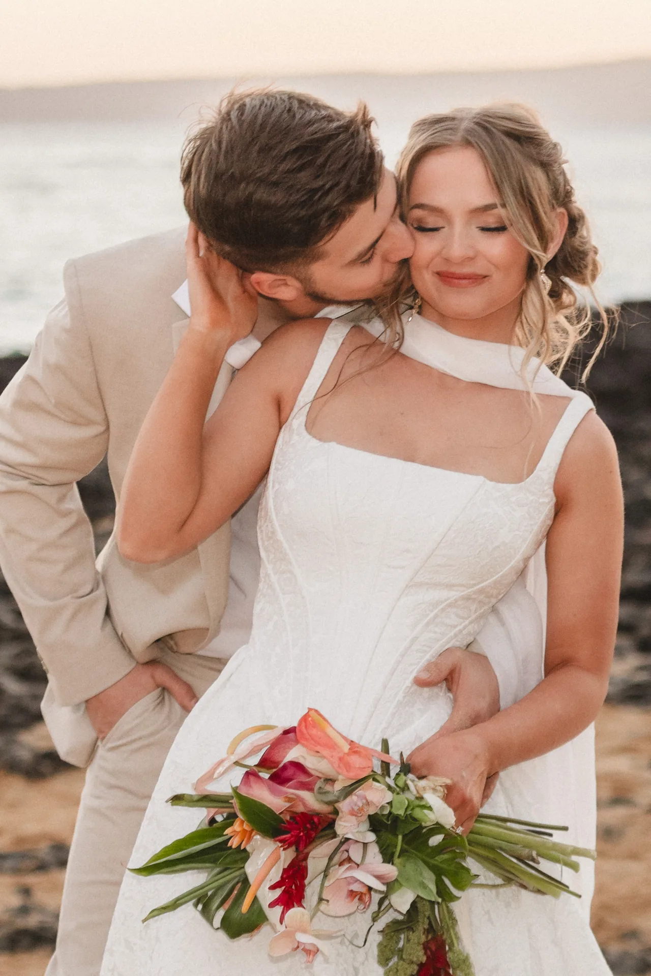 Groom in a tan suit kissing his smiling bride’s cheek as she holds a tropical bouquet of anthurium and orchids