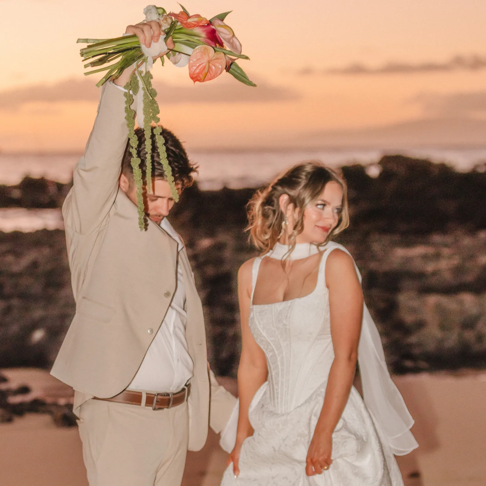 Groom playfully raising a tropical bouquet high in the air as his bride smiles beside him at sunset