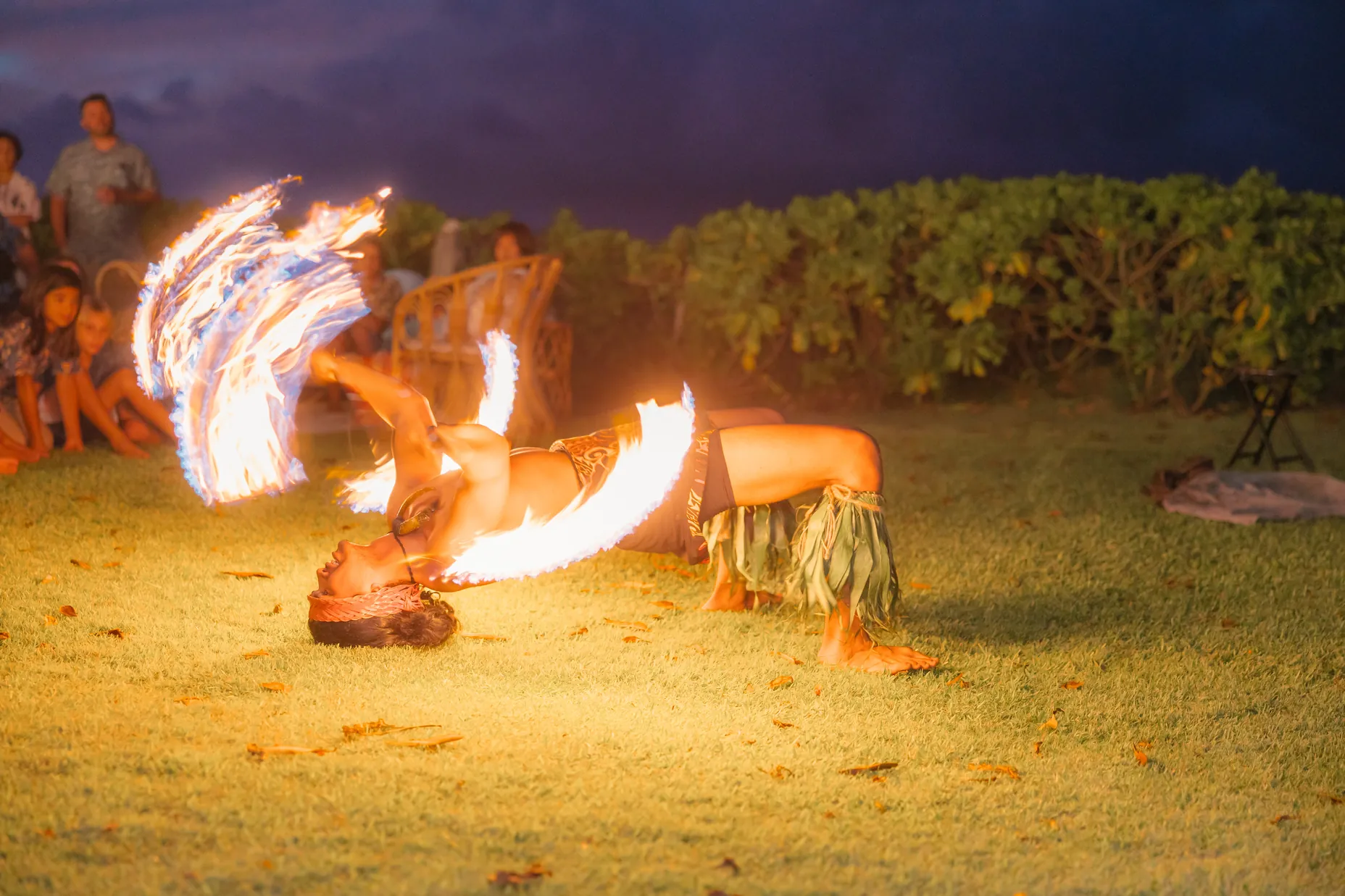 Fire knife dancer bending back on the lawn as spinning flames trace circles of light while guests watch