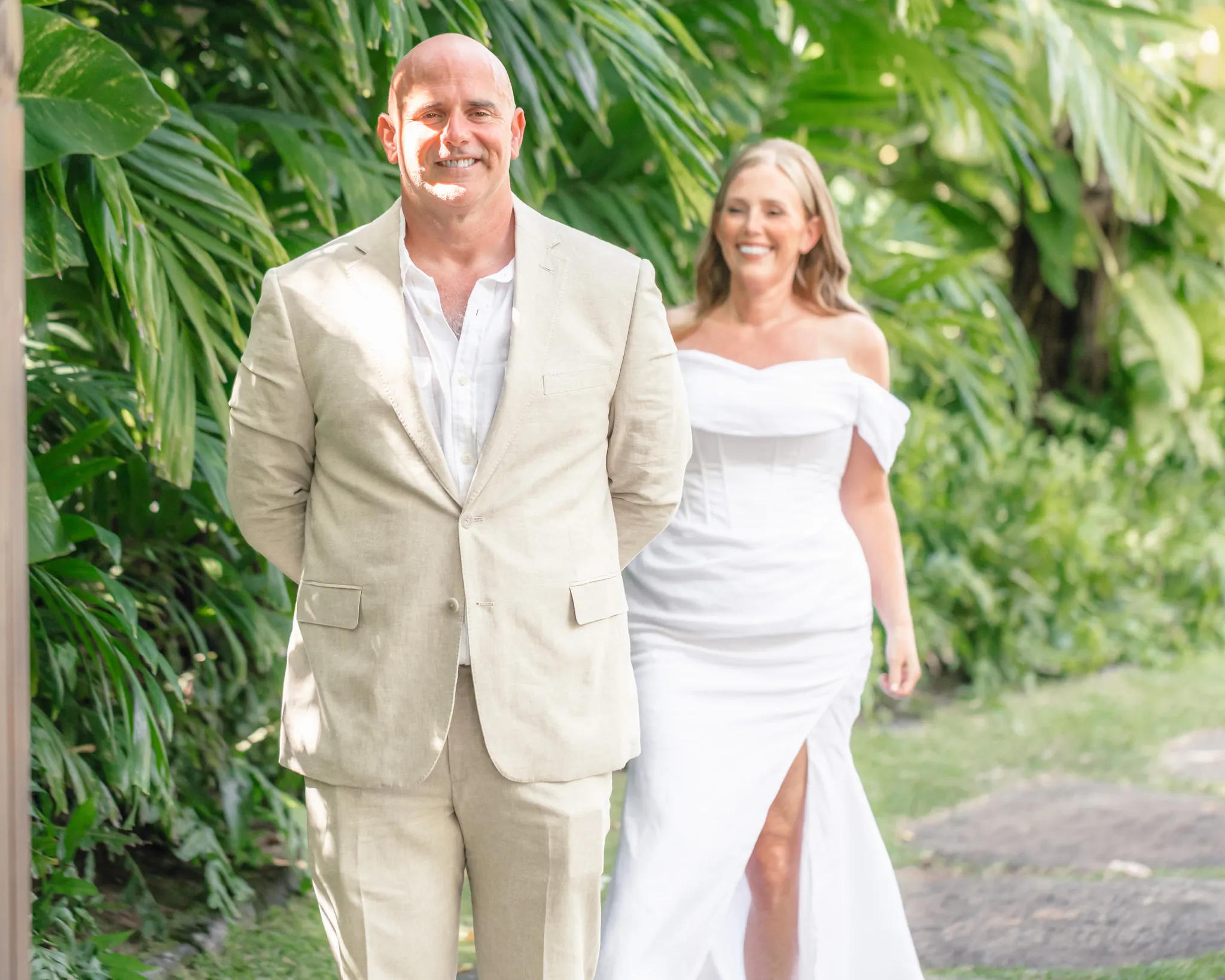 Groom in a linen suit smiling as his bride walks up beside him for their first look in a tropical garden