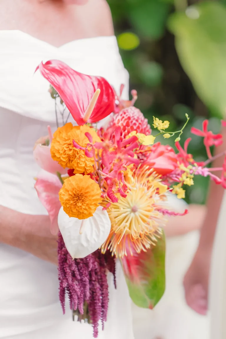 Bridal bouquet of pink anthurium, orange marigolds, protea and amaranthus held against a white wedding gown
