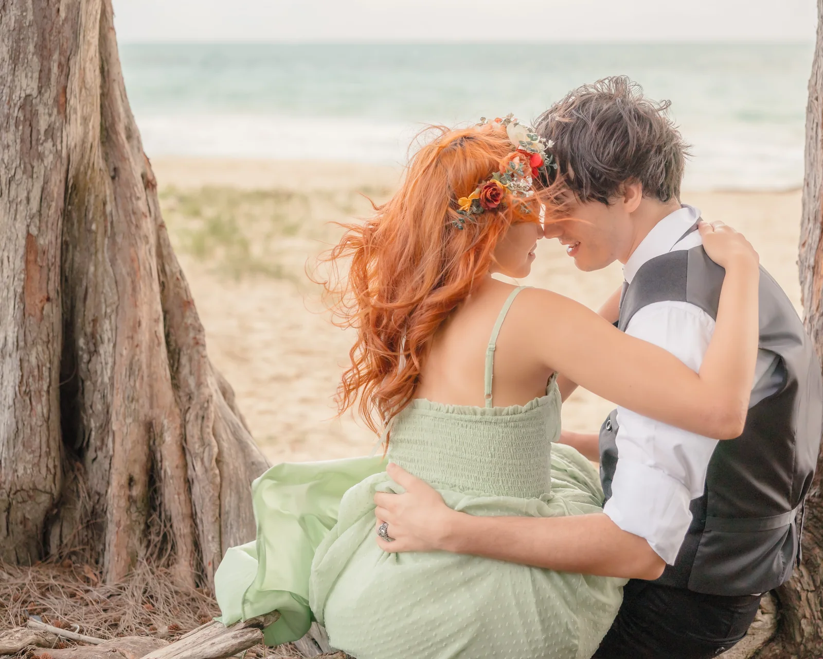 Woman with long red hair and a flower crown sitting with her partner beneath a tree on the beach, foreheads close together