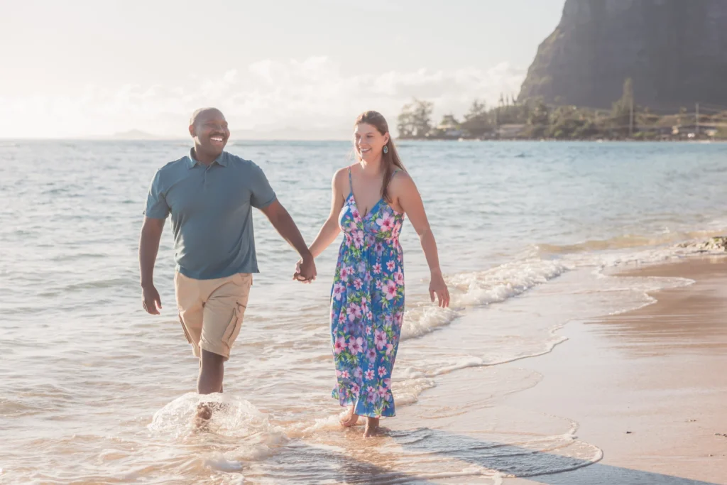 Engaged couple walking hand in hand through the shallows at Kaʻaʻawa Beach with the Koʻolau mountains behind them
