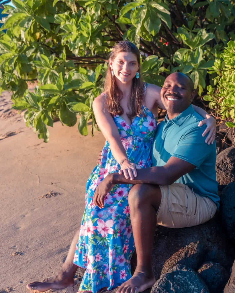 Engaged couple sitting close on the rocks in front of tropical greenery at Kaʻaʻawa Beach, Oahu