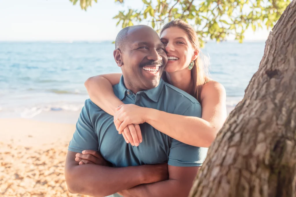 Groom-to-be laughing as his fiancée wraps her arms around him under a shade tree at the water's edge, Oahu