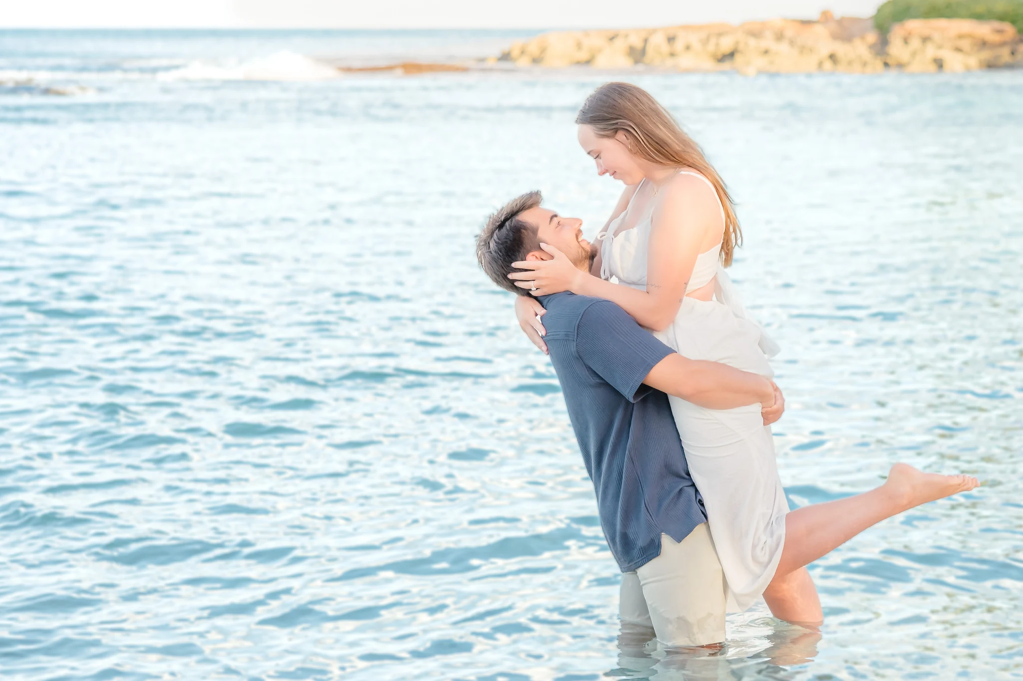 Man lifting his fiancée in clear turquoise water as she smiles down at him and kicks one foot up
