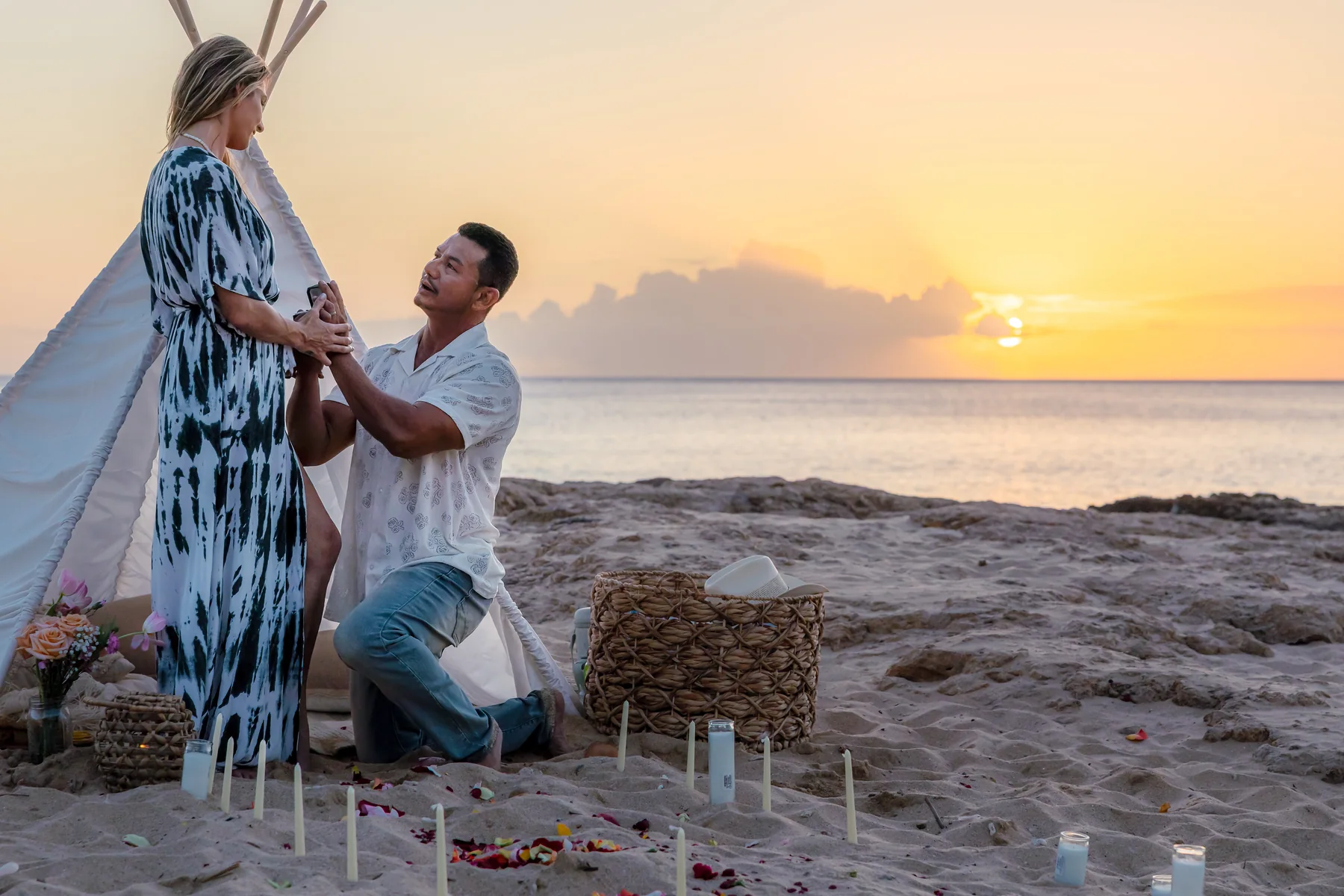 Man kneeling in the sand beside a white teepee and candles to propose as the sun sets over the ocean
