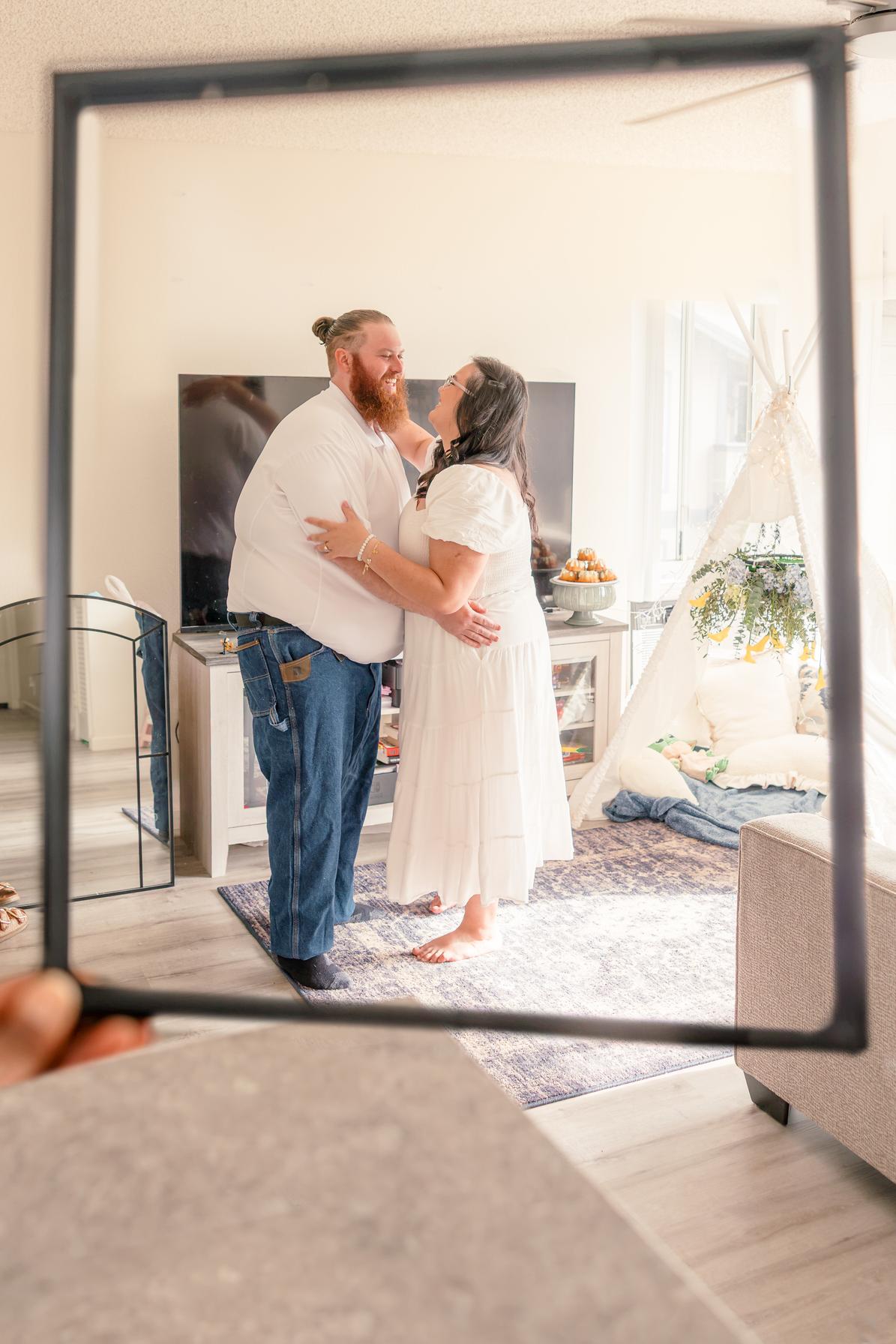 Hope and Dean embrace as they dance in their living room, seen through an empty picture frame.
